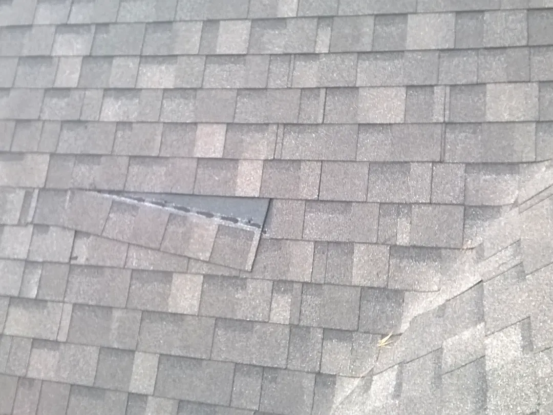 A grey shingle roof with one lifted, damaged shingle before repair