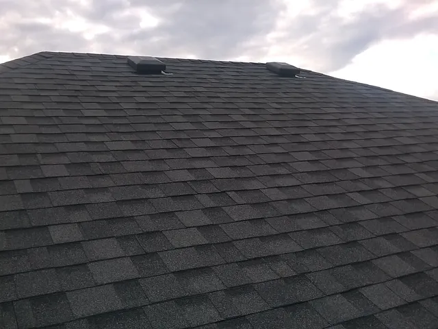 Dark shingle roof with two new roof vents against a cloudy sky